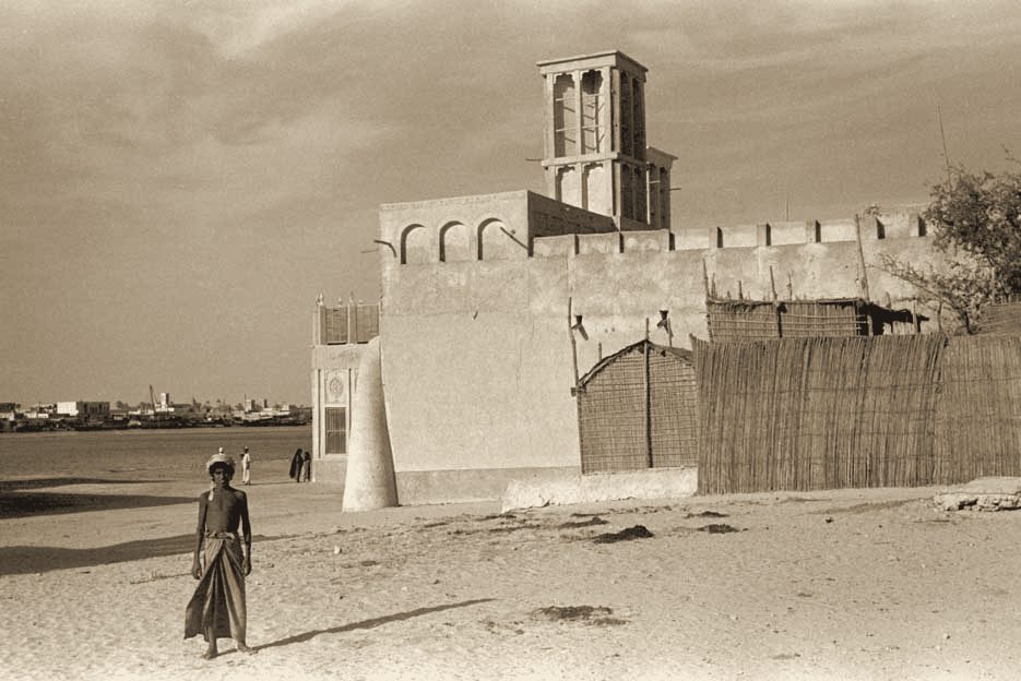A man walking across the sand in front of a wind-tower house, 1948