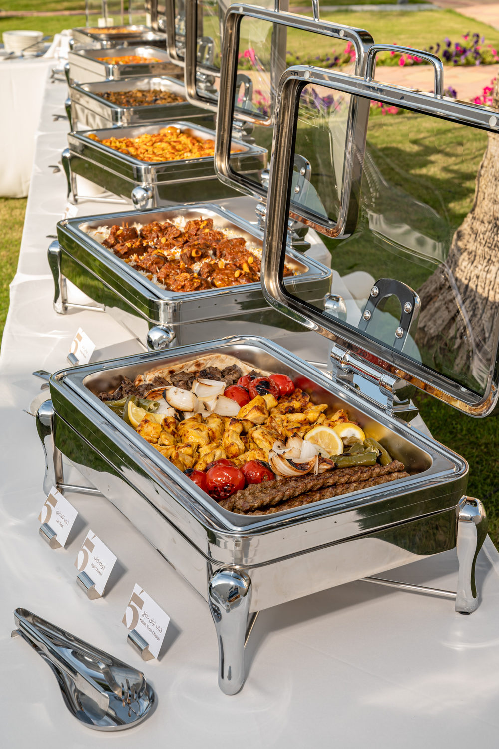 A Table 5 buffet of chafing dishes with grilled kebabs, chicken and rice, set up outdoors